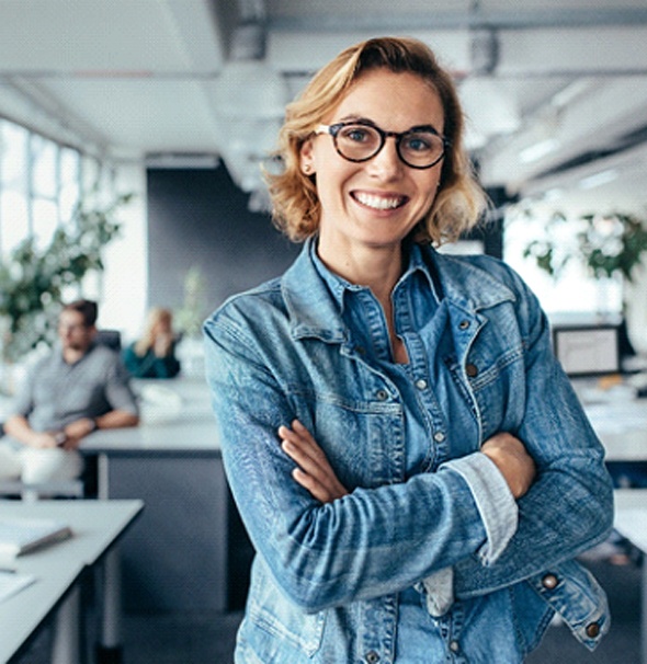 A middle-aged woman wearing glasses and a denim blouse while standing in an office and smiling after seeing her dentist in Braintree about a fixed bridge