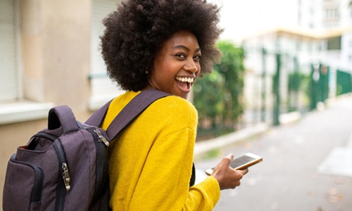 A young female wearing a yellow sweater, purple backpack, and holding her cell phone while smiling after receiving her new dental crown